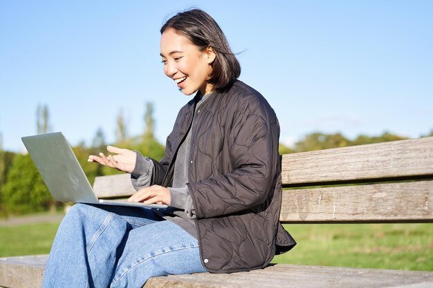 Kostenloses Foto porträt eines lächelnden asiatischen mädchens sitzt auf einer bank im park und spricht mit einem freund online über laptop-video-chats u