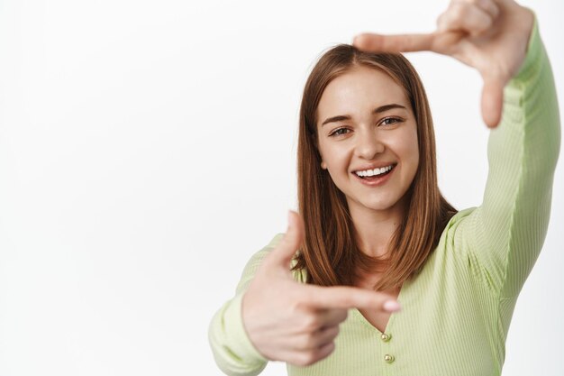 Porträt eines lächelnden blonden Mädchens, das durch kreative Fingerrahmen schaut, lacht, sich etw vorstellt, Kameraaufnahmen macht, glücklich vor weißem Hintergrund steht