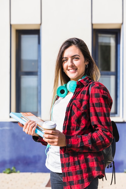 Porträt eines lächelnden Hochschulstudenten, der in der Hand Bücher und die Wegwerfkaffeetasse zur Kamera schaut hält