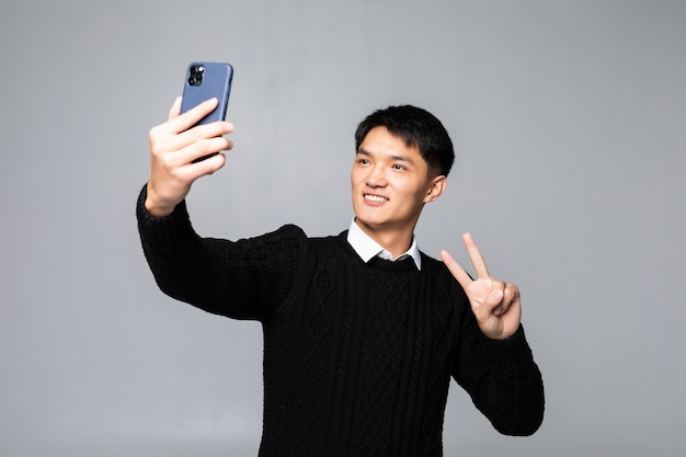 Porträt eines lächelnden jungen chinesischen Mannes, der ein Selfie mit Handy nimmt, während über weißer Wand lokalisiert