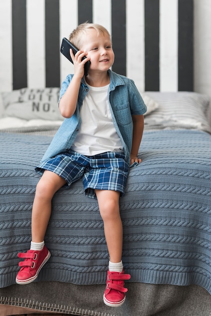 Porträt eines lächelnden Jungen, der auf dem Bett spricht am intelligenten Telefon sitzt