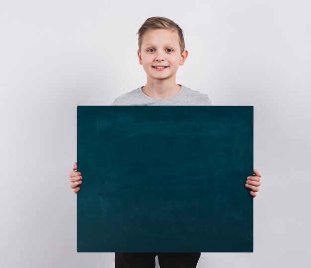 Porträt eines lächelnden Jungen, der leere Tafel gegen grauen Hintergrund hält