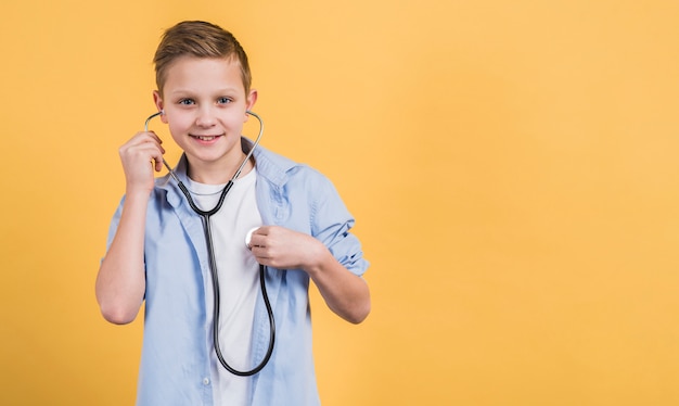 Porträt eines lächelnden Jungen, der seinen Herzschlag mit Stethoskop gegen gelben Hintergrund überprüft