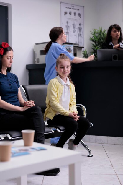 Porträt eines lächelnden jungen Mädchens mit ihrer Mutter, die in der Krankenhauslobby sitzt und darauf wartet, einen Kinderarzt zu sehen. Eltern mit Kind an der Rezeption der Privatklinik zu Besuch beim Kinderarzt.