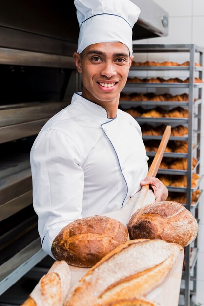 Porträt eines lächelnden jungen männlichen Bäckers, der frisch gebackenes Brot auf hölzerner Schaufel zeigt
