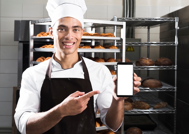 Kostenloses Foto porträt eines lächelnden jungen männlichen bäckers, der handy vor gebackenen hörnchenregalen zeigt