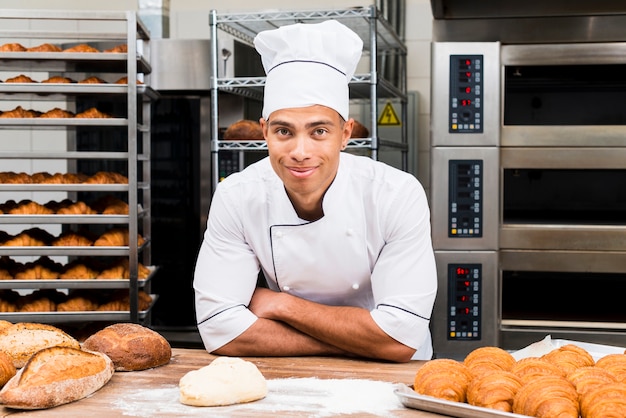 Porträt eines lächelnden jungen männlichen Bäckers, der hinter der Tabelle mit frischem Hörnchen und Brotlaib steht