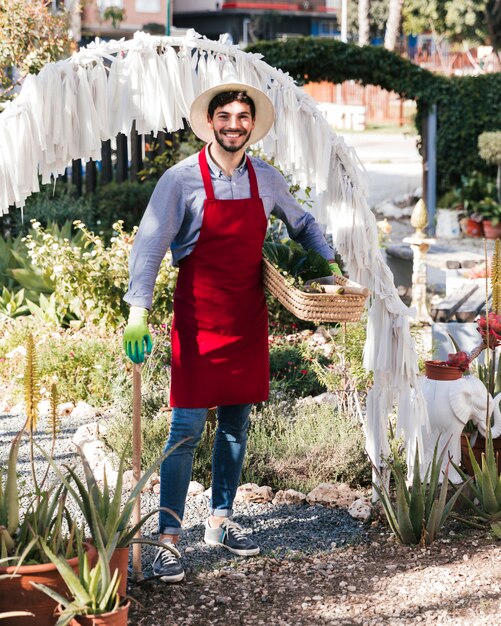 Porträt eines lächelnden jungen männlichen Gärtners, der Gartenarbeitwerkzeug und -korb hält