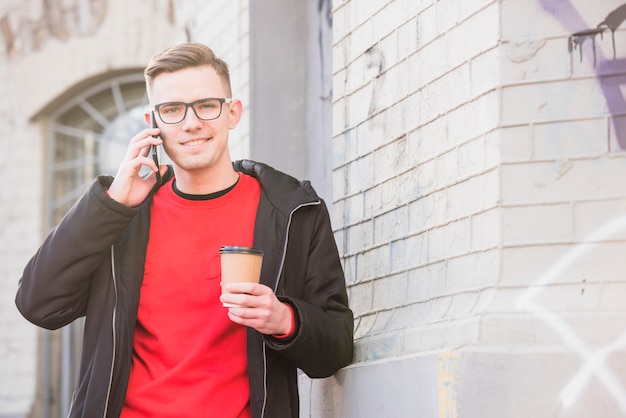 Porträt eines lächelnden jungen Mannes, der am Handy hält Mitnehmerkaffee spricht