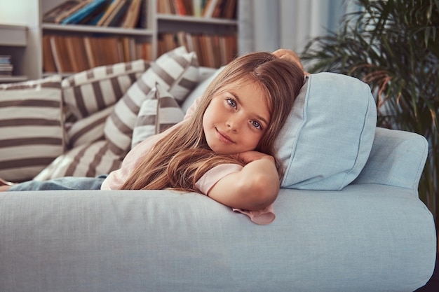 Porträt eines lächelnden kleinen Mädchens mit langen braunen Haaren und durchdringendem Blick, das zu Hause auf einem Sofa liegt.