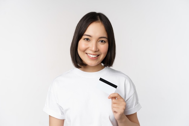 Porträt eines lächelnden koreanischen Bankkunden, der eine Kreditkarte mit einem glücklichen Gesicht zeigt, das vor weißem Hintergrund steht
