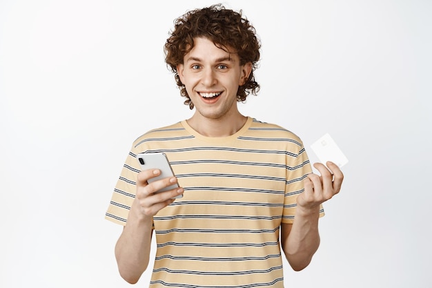 Porträt eines lächelnden, lockigen Kerls, der Kreditkarte und Smartphone mit App hält, um online über weißem Hintergrund einzukaufen