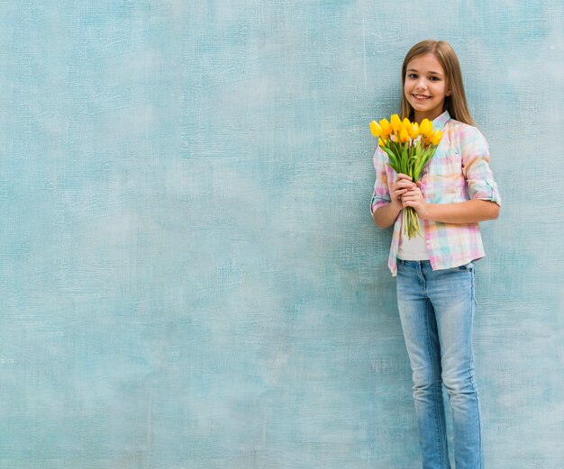 Porträt eines lächelnden Mädchens, das gelbe Tulpe hält, blüht in der Hand, die gegen blaue Wand steht