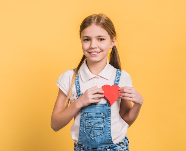 Porträt eines lächelnden Mädchens, das rotes Papier zeigt, schnitt Herzform gegen gelben Hintergrund heraus