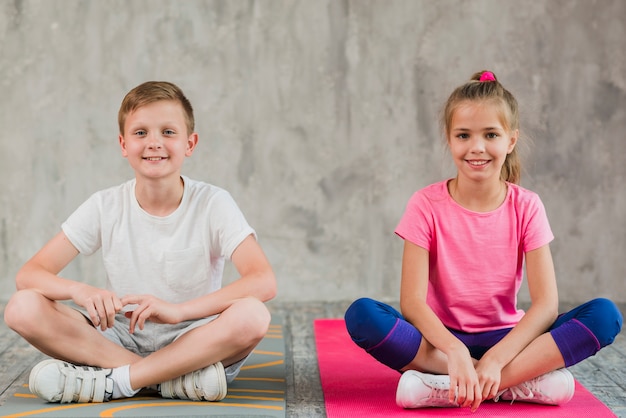 Porträt eines lächelnden Mädchens und des Jungen, die auf Übungsmatte mit ihren gekreuzten Beinen vor Wand sitzen