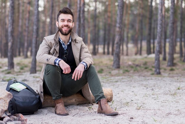 Porträt eines lächelnden männlichen Reisenden, der auf Strand mit seinem Rucksack sitzt