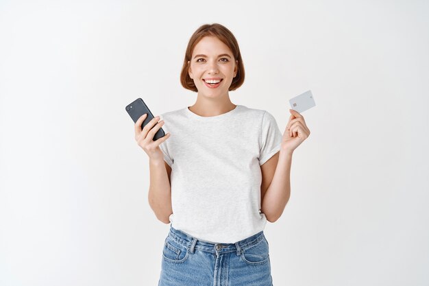 Porträt eines lächelnden natürlichen Mädchens, das Handy und Plastikkreditkarte zeigt, online bezahlt und gegen weiße Wand steht