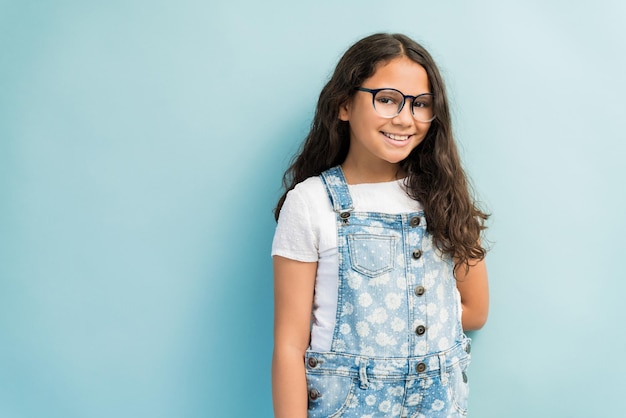 Porträt eines lächelnden vorpubertären Mädchens, das Denim-Overalls trägt, während es vor türkisfarbenem Hintergrund steht