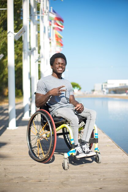 Porträt eines lässigen Mannes im Rollstuhl. Afroamerikanischer Mann in legerer Kleidung am Damm, Victory-Zeichen zeigend. Blauer Himmel und Fahnen im Hintergrund. Porträt, Schönheit, Glückskonzept