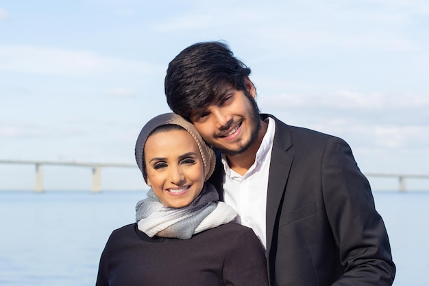Porträt eines liebevollen arabischen Paares während des Spaziergangs. Frau mit bedecktem Kopf und hellem Make-up und Mann im Anzug, der in die Kamera lächelt und sich umarmt. Liebe, Zuneigung Konzept
