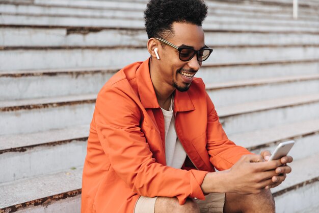 Porträt eines lockigen dunkelhäutigen Mannes in orangefarbener Jacke und Sonnenbrille, der aufrichtig lächelt und das Telefon hält