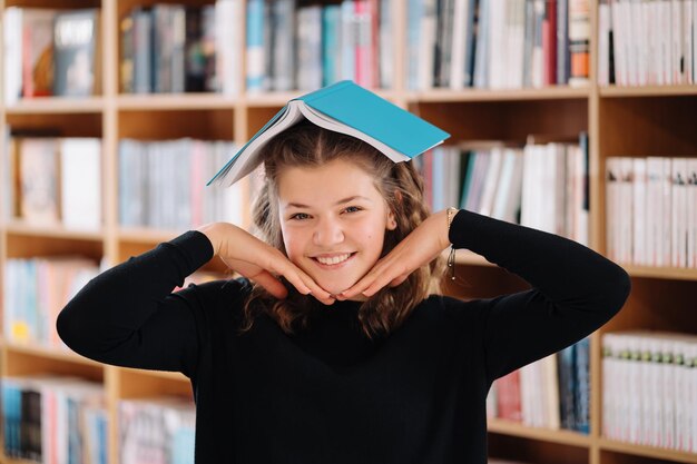 Porträt eines lustig lächelnden kaukasischen Teenagermädchens, das ein Buch auf den Kopf legt und hält und in die Kamera schaut, Bücherregalhintergrund.