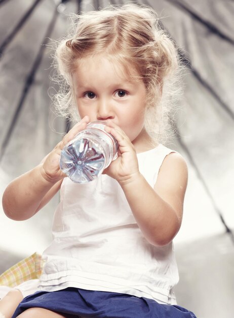 Porträt eines lustigen, süßen kleinen Mädchens in einem lässigen Kleid, trinkt Wasser, während es in einem Studio sitzt.