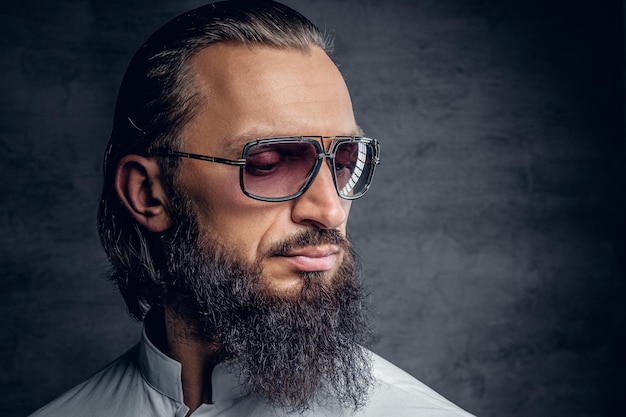 Porträt eines luxuriösen bärtigen Mannes mit Sonnenbrille, gekleidet in weißem Hemd auf grauem Hintergrund.