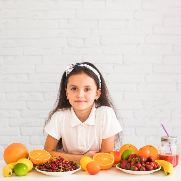 Porträt eines Mädchens, das auf Schreibtisch mit vielen verschiedenen Früchten sich lehnt