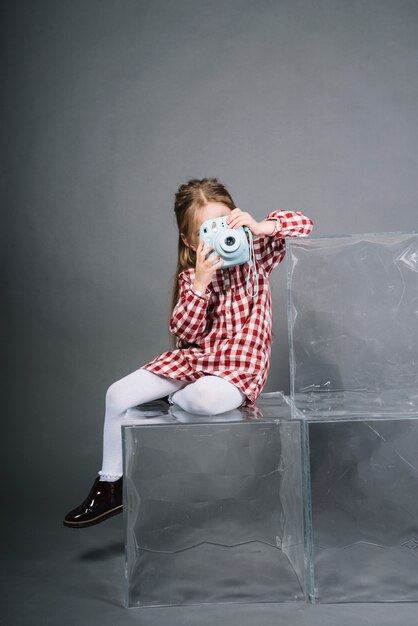Porträt eines Mädchens, das durch die sofortige Kamera sitzt auf transparenten Würfeln gegen grauen Hintergrund fotografiert