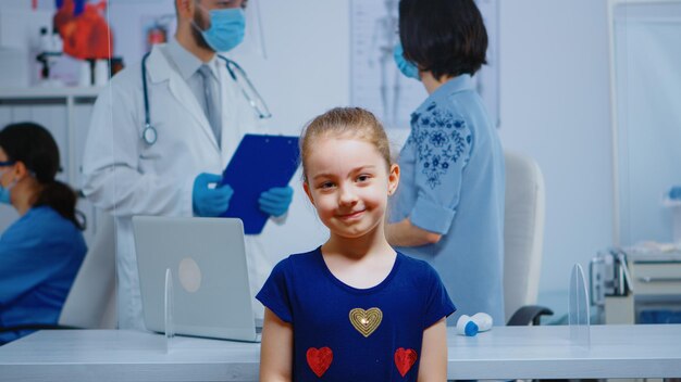 Porträt eines Mädchens, das in der Arztpraxis lächelt, während die Mutter mit dem Arzt im Hintergrund spricht. Facharzt für Medizin mit Schutzmaske im Gesundheitswesen, Beratung in Krankenhausklinik