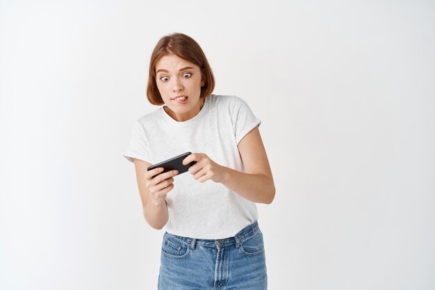 Porträt eines Mädchens, das sich auf Videospiele konzentriert, auf dem Smartphone spielt und auf die Lippe beißt, den Körper mit dem Handy kippt, weiße Wand