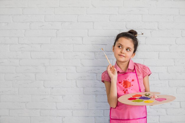 Porträt eines Mädchens mit dem Malerpinsel und Palette, die gegen weiße Wand stehen