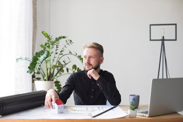 Porträt eines männlichen Architekten mit Hausmodell auf Architektenplan am Arbeitsplatz