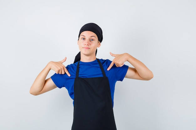 Porträt eines männlichen Teenagerkochs, der in T-Shirt, Schürze auf sich selbst zeigt und selbstbewusste Vorderansicht schaut