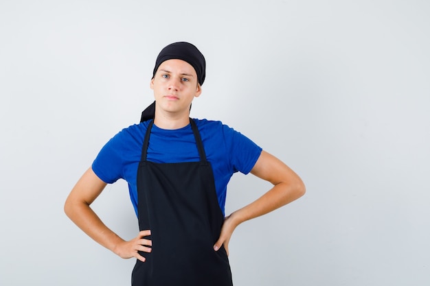 Kostenloses Foto porträt eines männlichen teenagerkochs mit den händen an der taille in t-shirt, schürze und selbstbewusster vorderansicht