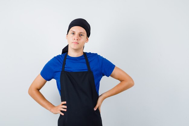 Porträt eines männlichen Teenagerkochs mit den Händen an der Taille in T-Shirt, Schürze und selbstbewusster Vorderansicht