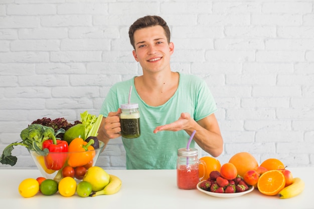 Porträt eines Mannes, der grünes Smoothieflasche mit vielen gesundem Lebensmittel auf Tabelle hält