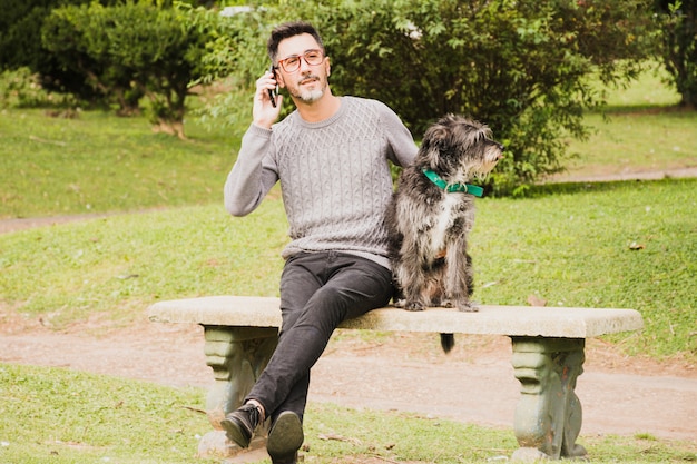 Porträt eines modernen Mannes, der im Park mit seinem Hund spricht am Handy sitzt
