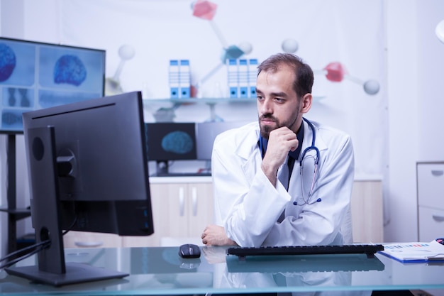 Porträt eines nachdenklichen jungen Arztes, der den Monitor in seinem Kabinett untersucht. Doktor, der seinen Computer für die Arbeit verwendet.