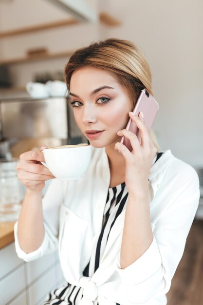 Porträt eines netten Mädchens, das in die Kamera schaut, während es an der Theke sitzt und Kaffee mit Handy in der Hand im Café trinkt. Hübsche Dame mit blonden Haaren, die im Café auf ihrem Handy spricht