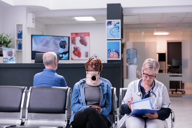 Porträt eines Patienten mit Halskragenschaum, der im Wartezimmer einer Klinik sitzt und sich mit einem leitenden Arzt berät. Frau mit Halskrause nach Unfallverletzung im Gespräch mit Sanitäter über Gesundheitsversorgung.
