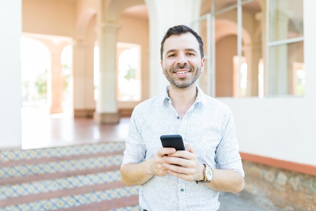 Porträt eines positiven mittleren erwachsenen Mannes, der lächelt, während er sein Handy gegen das Gebäude hält