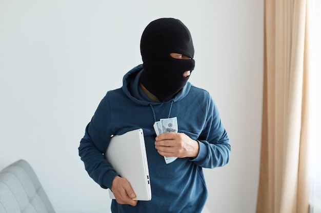 Porträt eines räubers mit blauem hoodie und schwarzer sturmhaube, der laptop und bargeld im schlauch einer person ausraubt, den ort des verbrechens verlässt, das fenster ansieht, illegale handlungen. Premium Fotos