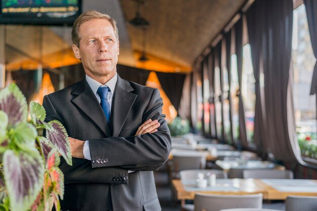 Porträt eines reifen Geschäftsmannes mit den gekreuzten Armen, die im Restaurant stehen