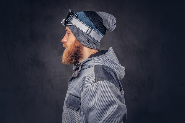 Kostenloses Foto porträt eines rothaarigen snowboarders mit vollbart in wintermütze und schutzbrille in einem snowboardmantel, der in einem studio posiert und wegschaut. getrennt auf einem dunklen strukturierten hintergrund.