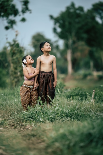 Porträt eines schönen asiatischen Jungen mit nacktem Oberkörper und Mädchen in thailändischer traditioneller Kleidung und schöne Blume auf ihr Ohr, Hand in Hand stehend und mit lächelndem Kopienraum in den Himmel schauend