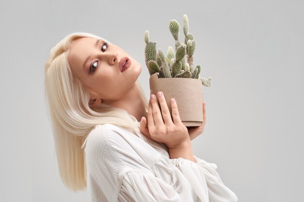 Porträt eines schönen blonden Mädchens mit Make-up und Frisur, das weiße Bluse trägt, in die Kamera schaut und Topf mit grünem Kaktus hält. Hübsche junge Frau kauft Pflanze für Haus