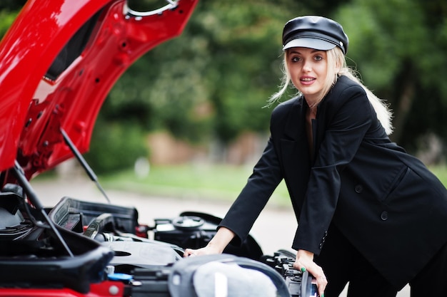 Porträt eines schönen blonden sexy Modefrauenmodells in Mütze und ganz in Schwarz mit hellem Make-up in der Nähe eines roten Stadtautos mit offener Motorhaube