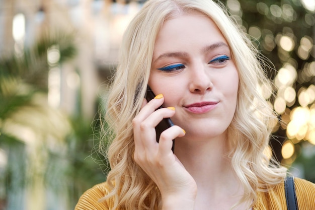Porträt eines schönen, flirty blonden Mädchens, das schlau wegschaut, während es auf dem Handy im Freien spricht